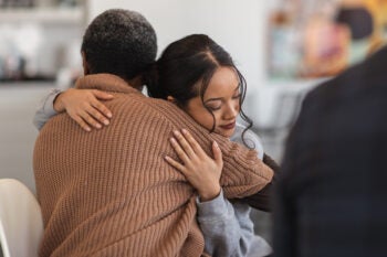 Two people embrace while seated, with a blurred person in the foreground.