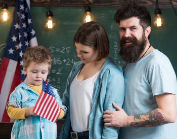 Kid with parents in classroom with usa flags, chalkboard on background. American family and cute son stand with usa flags. Patriotic education concept. Parents teaching son american traditions playing