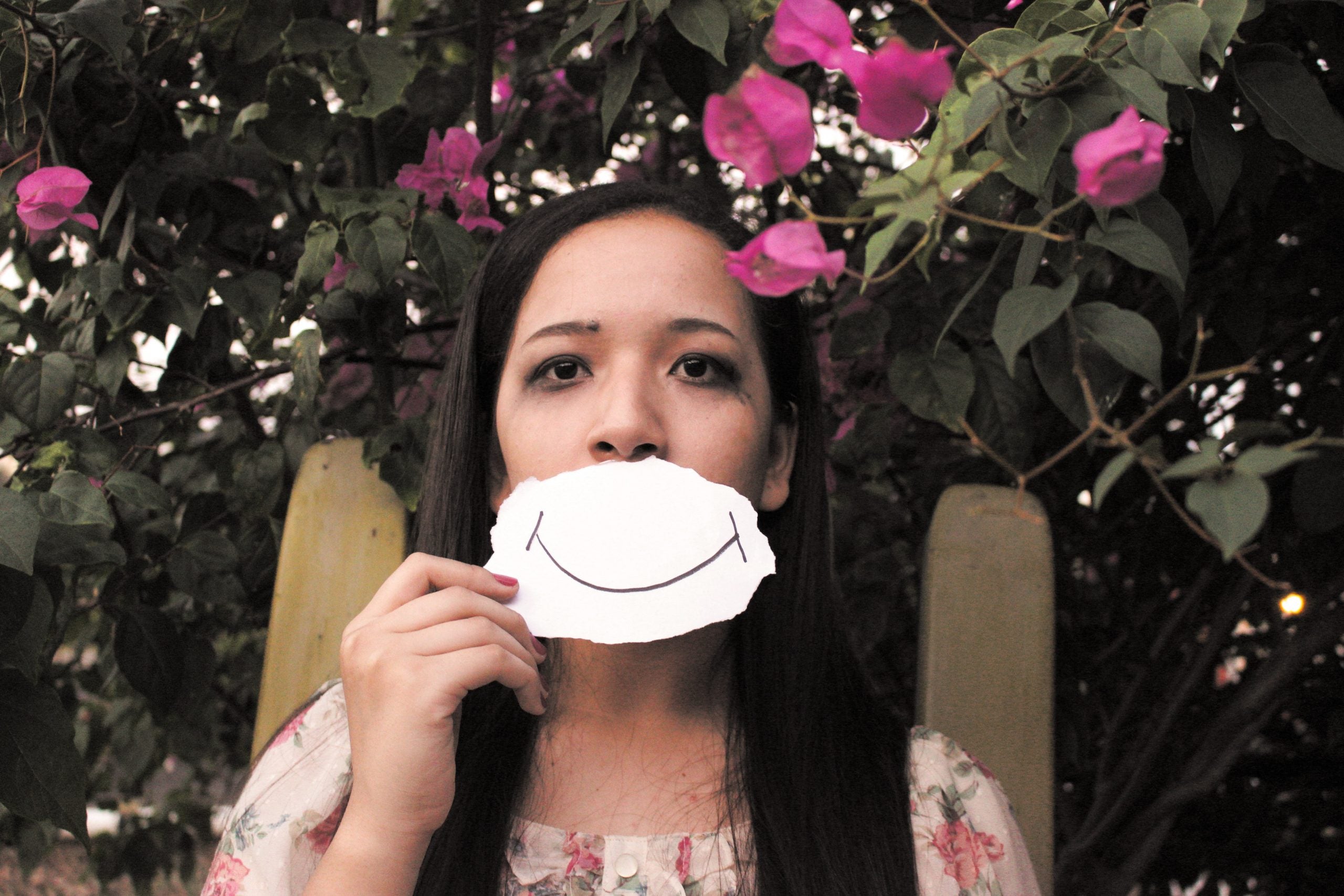 sad woman standing under pink petaled flowers holding up a smile drawing on a piece of paper