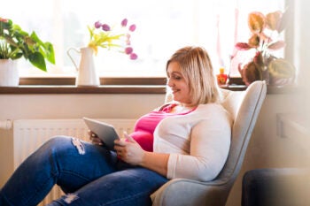 Attractive overweight woman at home sitting in armchair working on tablet.
