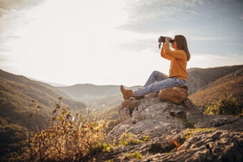 Woman with binoculars sitting on mountain peak in sunset